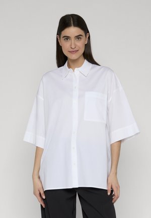 Junge Frau mit langen dunklen Haaren, die ein lockeres weißes Hemd mit Brusttasche und schwarze Hose trägt, vor einem schlichten Hintergrund stehend.