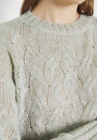 Pull en tricot vert clair avec un motif en torsades et un col rond. Le tissu a une texture douce et légère avec un léger éclat.