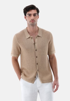 Junger Mann trägt ein beigefarbenes, kurzärmliges, gestricktes Hemd mit Knöpfen und Kragen sowie weiße Hosen, die Hände teilweise in den Taschen, neutraler Hintergrund.