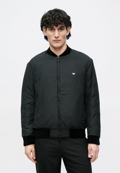 Μαύρο μπουφάν τύπου bomber με λείο ύφασμα, χρυσό λογότυπο στο στήθος, ριμπ γιακά και μανσέτες, καθώς και μπροστινό φερμουάρ. Απλός, λιτός σχεδιασμός.