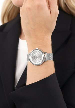 Silberne Uhr mit einem Mesh-Armband, runder Gehäuse und einem gemusterten hellgrauen Zifferblatt. Der Arm ist nackt und trägt ein schwarzes Blazer.