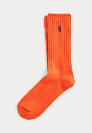 Calcetín de caña con canalé de color naranja brillante con logo bordado de jugador de polo en azul cerca del puño sobre un fondo blanco.