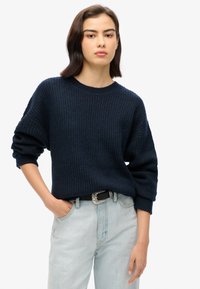 Dark navy knitted sweater with a round neckline and drop shoulder design, paired with light blue jeans and a silver buckle belt.