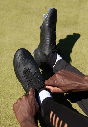 Person zieht schwarze Fußballschuhe fest, während sie auf Gras sitzt, trägt schwarze Hose und weiße Socken.
