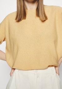 Żółta bluzka z krótkim rękawem wykonana z miękkiego materiału o luźnym kroju. Charakteryzuje się subtelną fakturą i okrągłym dekoltem, zestawiona z jasnymi spodniami.