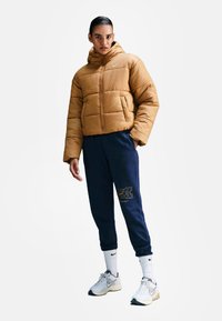 Brązowa kurtka ocieplana z kapturem, granatowe dresy z detalem logo, białe skarpetki i szare sneakersy. Model stoi na jednolitym tle.