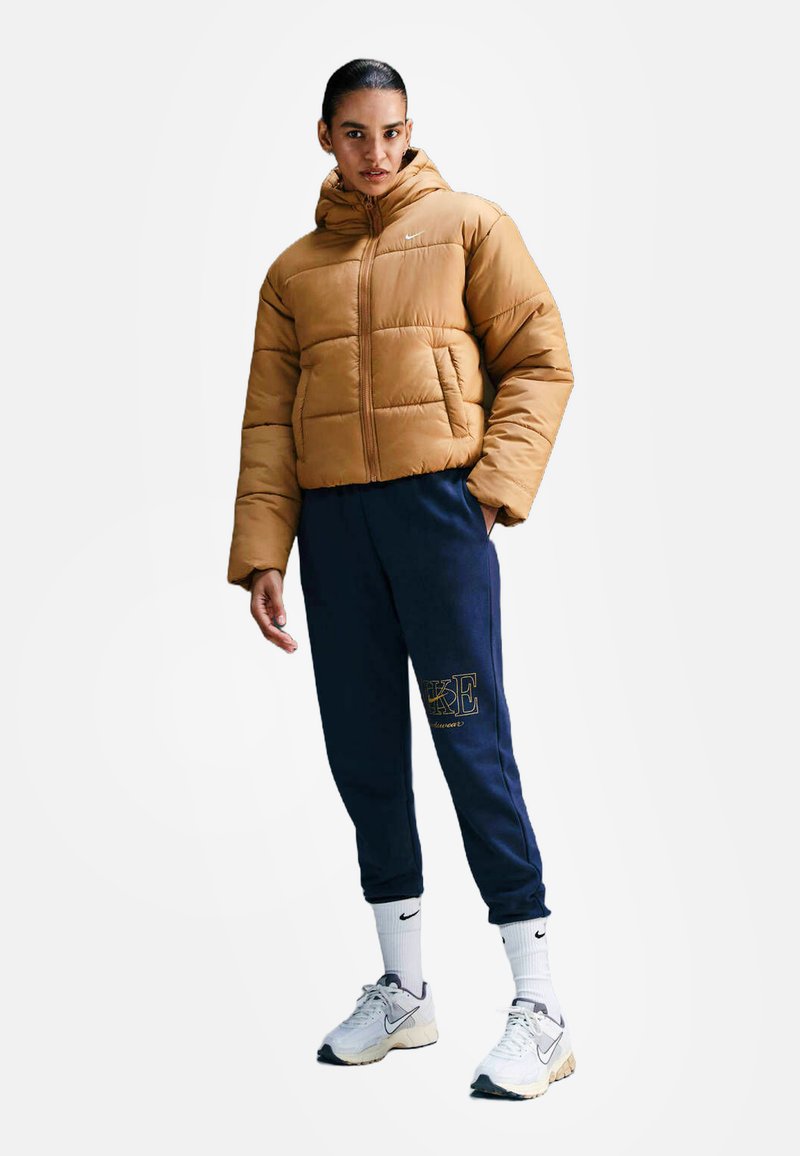 Brązowa kurtka ocieplana z kapturem, granatowe dresy z detalem logo, białe skarpetki i szare sneakersy. Model stoi na jednolitym tle.