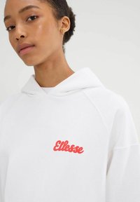 Młoda kobieta z krótkimi kręconymi włosami, ubrana w białą bluzę z kapturem z czerwonym logo "Ellesse" na piersi oraz małymi złotymi kolczykami w kształcie kółka.