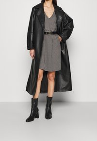 Svart läder trenchcoat med vida ärmar, bärs över en klänning med svart och vit houndstooth-mönster, åtsnörd med ett svart bälte och guldspänne.