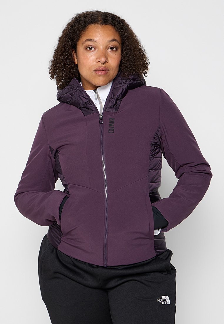 Donna che indossa una giacca con cappuccio viola Colmar e pantaloni neri The North Face, con le mani nelle tasche della giacca, in piedi contro uno sfondo chiaro e semplice.