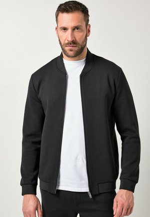 Mann trägt eine schwarze Jacke mit Reißverschluss über einem weißen T-Shirt, steht vor einem schlichten hellen Hintergrund.
