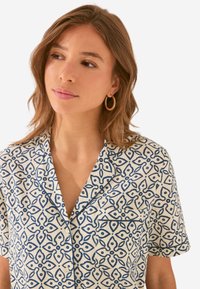 Korte mouwen blouse van lichte stof. Heeft een witte achtergrond met een blauw geometrisch patroon en een klein borstzakje.
