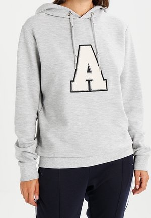Personne portant un sweat à capuche gris clair avec une grande lettre A blanche et noire à l'avant, assorti à un pantalon foncé.