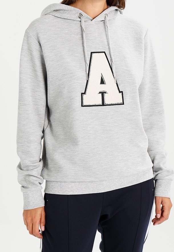 Personne portant un sweat à capuche gris clair avec une grande lettre A blanche et noire à l'avant, assorti à un pantalon foncé.