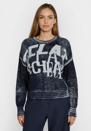 Frau trägt einen marineblauen und grauen Strickpullover mit großen, verzerrten weißen Buchstaben und marineblaue Hose, steht vor einem schlichten weißen Hintergrund.