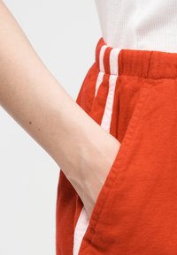 Χέρι που είναι βαλμένο στην πλαϊνή τσέπη κόκκινου παντελονιού με λευκές κάθετες ρίγες, φορώντας λευκό ribbed τοπ.