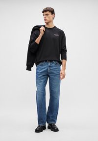 Svart langermet t-skjorte med logo, blå rette jeans og svarte lær-sko. Modellen har en svart jakke over skulderen.