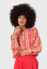 Blusa a scacchi arancione, rosa e gialla con maniche a sbuffo e chiusura con bottoni. Abbinata a pantaloni rossi a vita alta.