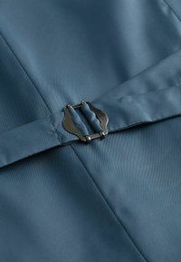 Un tissu de couleur bleu pâle présente une texture lisse. Un fermoir métallique est visible, maintenant un pli dans le matériau.