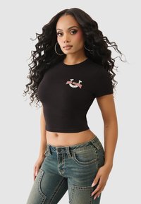 Czarny krótki t-shirt z okrągłym dekoltem, ozdobiony białym i czerwonym haftowanym logo, w zestawieniu z niebieskimi dżinsami.