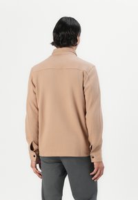 Homme de dos portant une chemise beige à manches longues avec poignets boutonnés et un pantalon gris foncé sur un fond blanc uni.
