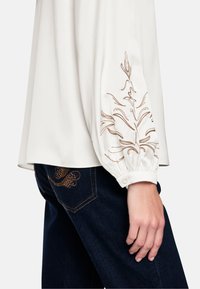 White blouse with gathered sleeves featuring gold embroidery along the outer arm, paired with dark denim jeans showing decorative stitching.