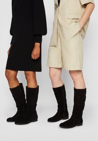 Dos mujeres que llevan botas de ante negro hasta la rodilla, una en un vestido negro y la otra en pantalones cortos beige y una chaqueta a juego.