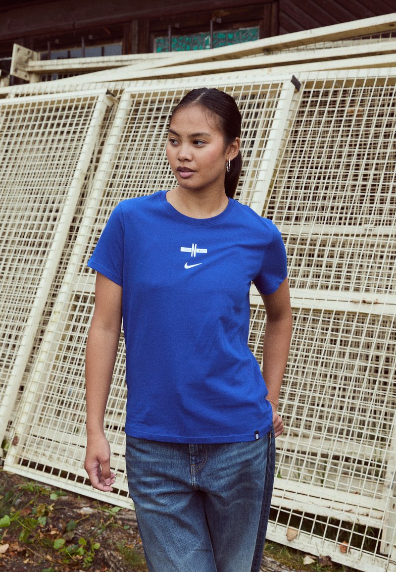 Mėlynas medvilninis t-shirt su apvalia iškirpte, ant kurio yra baltas Nike logotipas ir grafikos dizainas. Derinamas su šviesiai mėlynomis džinsinėmis kelnėmis.
