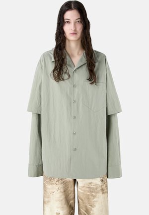 Pitkät tummahiuksinen henkilö pitää päällään ylisuurta vaaleanharmaata nappipaitaa ja beigenvärisiä, kuvioituja housuja yksinkertaista valkoista taustaa vasten.