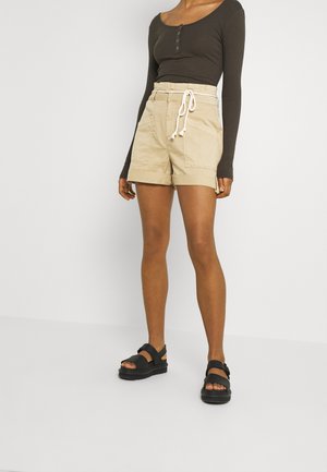 Person iført beige shorts med høj talje og opbukket kant med forlommer og hvid snor, kombineret med en mørk langærmet top og sorte sandaler.