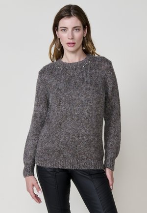Suéter gris de punto con un acabado texturizado, cuello redondo y detalles decorativos de cuentas en el collar. Llevado con pantalones de cuero negros.