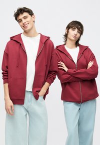 Bordó cipzáras kapucnis pulóverek elülső zsebekkel, fehér pólók fölött viselve. Puha anyag, laza szabás, világoskék bő szárú farmerral párosítva.