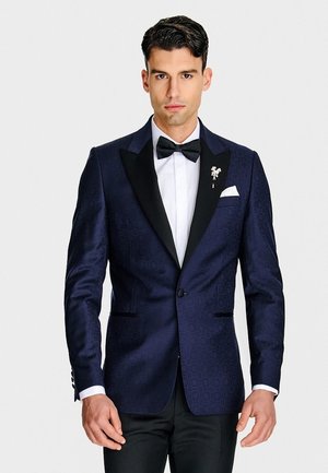 Mężczyzna w ciemnoniebieskiej marynarce smokingowej z czarnymi klapami, białej koszuli, czarnej muszce i białej poszetce, stojący na białym tle.