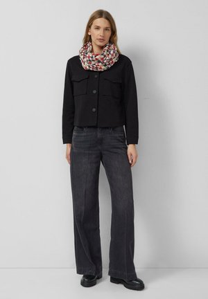 Frau steht, trägt eine schwarze, geknöpfte Jacke, weite dunkle Jeans, schwarze Schuhe und einen gemusterten roten, weißen und schwarzen Schal um den Hals.