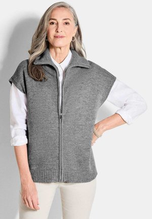 Gilet en maille grise avec col montant et manches courtes, doté d'une fermeture éclair sur le devant. Porté sur une chemise blanche, avec un bord côtes en détail.