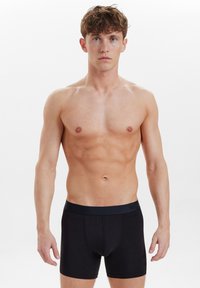 Musta puuvillainen boxeri, jossa on sileä pinta ja tumma vyötärö. Malli seisoo neutraalissa asennossa valkoista taustaa vasten.