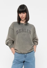 Felpa verde oliva con la scritta "BERLIN" stampata sul petto. Presenta polsini e colletto a coste, una texture morbida e una vestibilità rilassata.