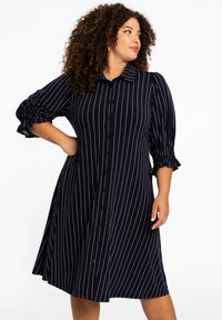 Knielanges Kleid in Marineblau mit weißen vertikalen Streifen. Verfügt über einen Kragen und gepuffte Ärmel mit Raffung. Glatte Stofftextur.