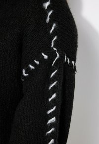 Schwarzer Pullover mit strukturiertem Strickmuster, der weiße Nähte entlang der Ärmel für Kontrast und Detailierung aufweist.