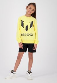 Felpa gialla con grafica nera, "MESSi," abbinata a pantaloni corti neri e sneakers bianche con le cavigliere e dettagli. 