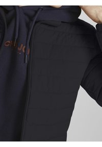 Svart puffjacka med quiltat mönster och dragkedjestängning, lager-på-lager över en marinblå huva med tydlig orange logotyp. Slät textur.