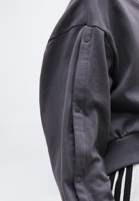 Tmavosivý rukáv mikiny s patentkami pozdĺž vonkajšieho švu na ramene na jednofarebnom pozadí.