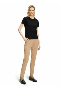 Czarna koszulka z krótkim rękawem połączona z beżowymi spodnimi spodniami o tapered fit. Modelka stoi z jedną ręką w kieszeni, nosząc czarne mokasyny. Czysty, minimalistyczny design.