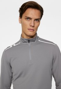 Grå långärmad zip-up skjorta med hög krage, med vita detaljer på axlarna och en texturerad tyg.