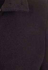 Zwarte stof met een gestructureerde afwerking en een kraagontwerp. Zichtbare stiksels bij de halslijn en details aan de zijnnaad.