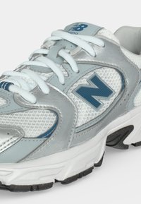 Λευκό και ασημί παπούτσι τρεξίματος New Balance με μπλε λογότυπο, λευκά κορδόνια και υφασμάτινη δίχτυ ψηφιδωτή υφή στο επάνω μέρος.