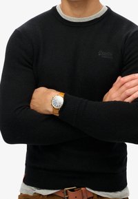 Maglione nero lavorato a maglia con scollo rotondo grigio, caratterizzato da un piccolo logo. Un orologio marrone con quadrante blu è indossato al polso.