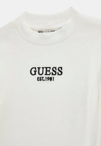 Pull long à manches longues blanc à côtes avec le logo "GUESS" brodé en noir et le texte "EST.1981" sur le devant. Design classique à col rond.
