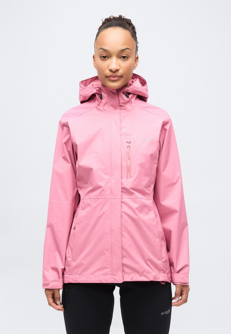 Νέα γυναίκα που φοράει ροζ αδιάβροχο μπουφάν με κουκούλα και τσέπες με φερμουάρ, στέκεται όρθια μπροστά σε απλό φωτεινό φόντο.