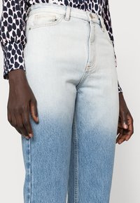 Jeans de ganga azul claro com um efeito ombré, transicionando de tons claros para mais escuros na parte inferior. Cintura alta com bolsos laterais.